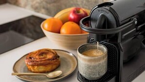 Uma cozinha com máquina de café, uma tigela de frutas, um prato com um doce e uma xícara de café.