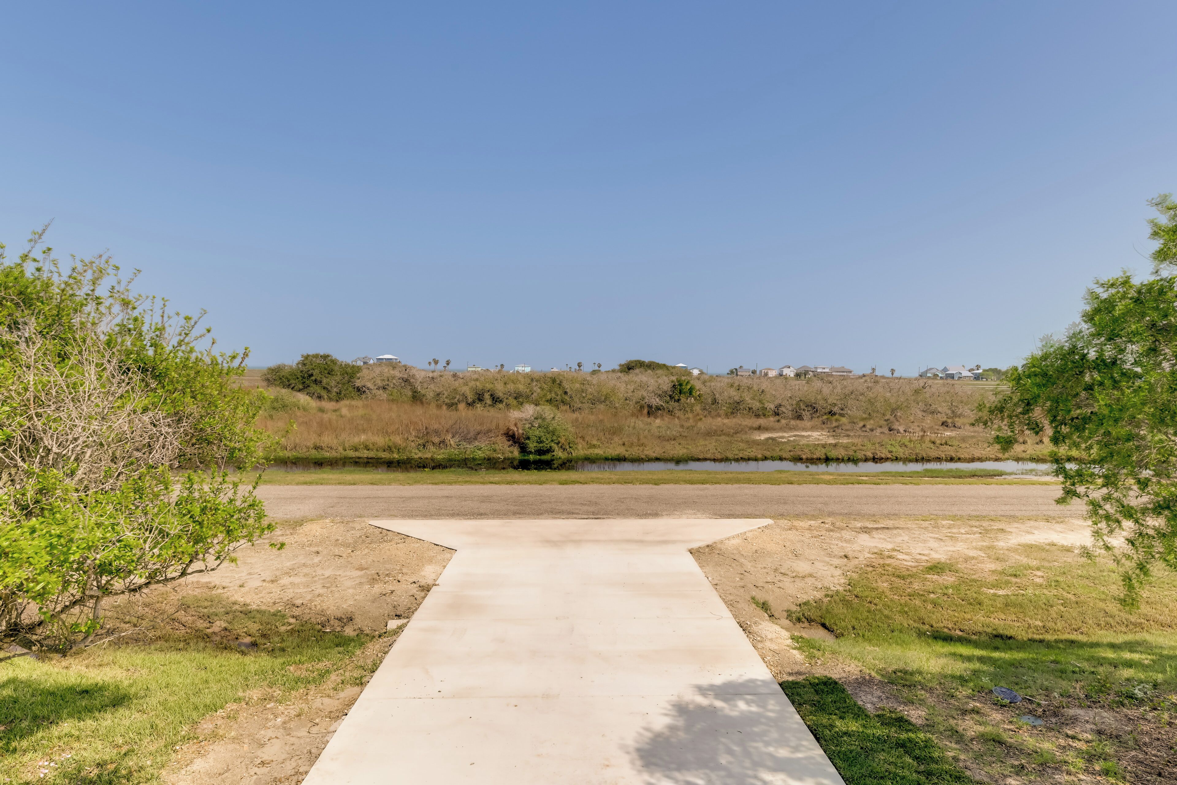 Um caminho de concreto leva a um corpo d'água, com vegetação escassa e edifícios ao longe.