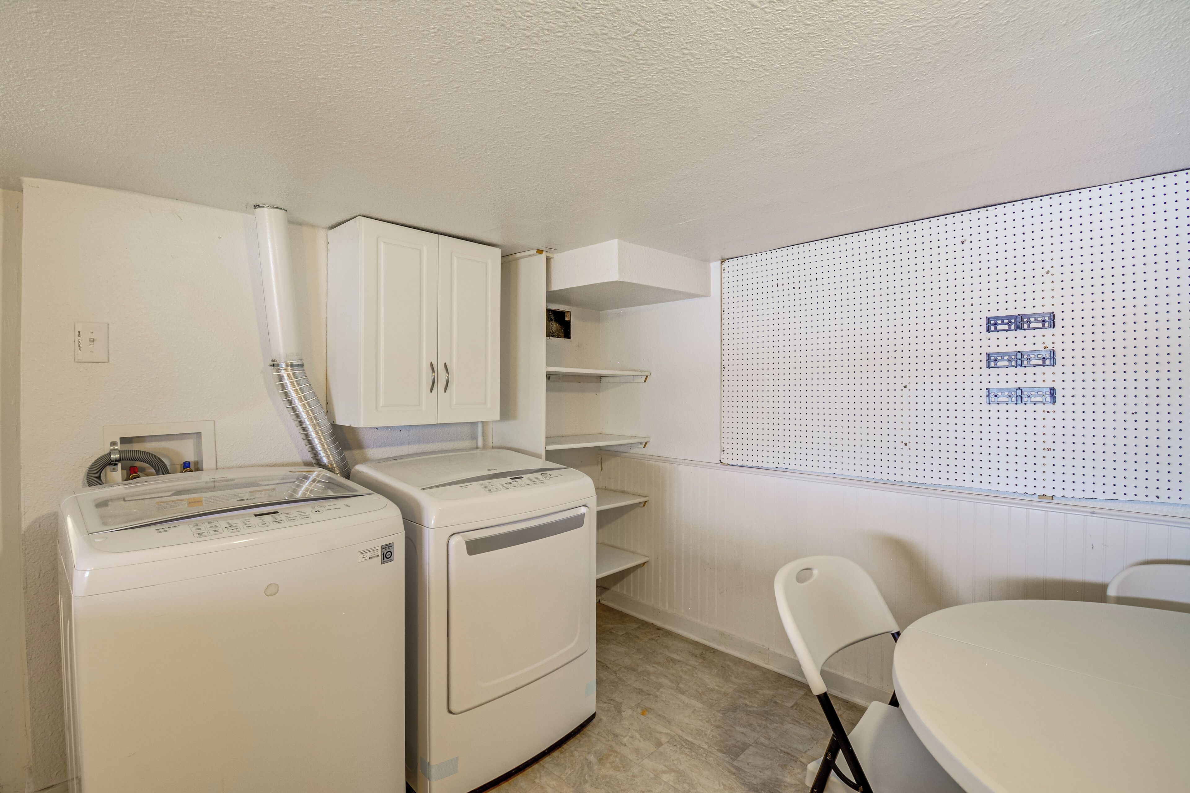 Uma lavanderia compacta com máquina de lavar e secadora de abertura frontal, painel de ganchos e uma pequena mesa com cadeiras.
