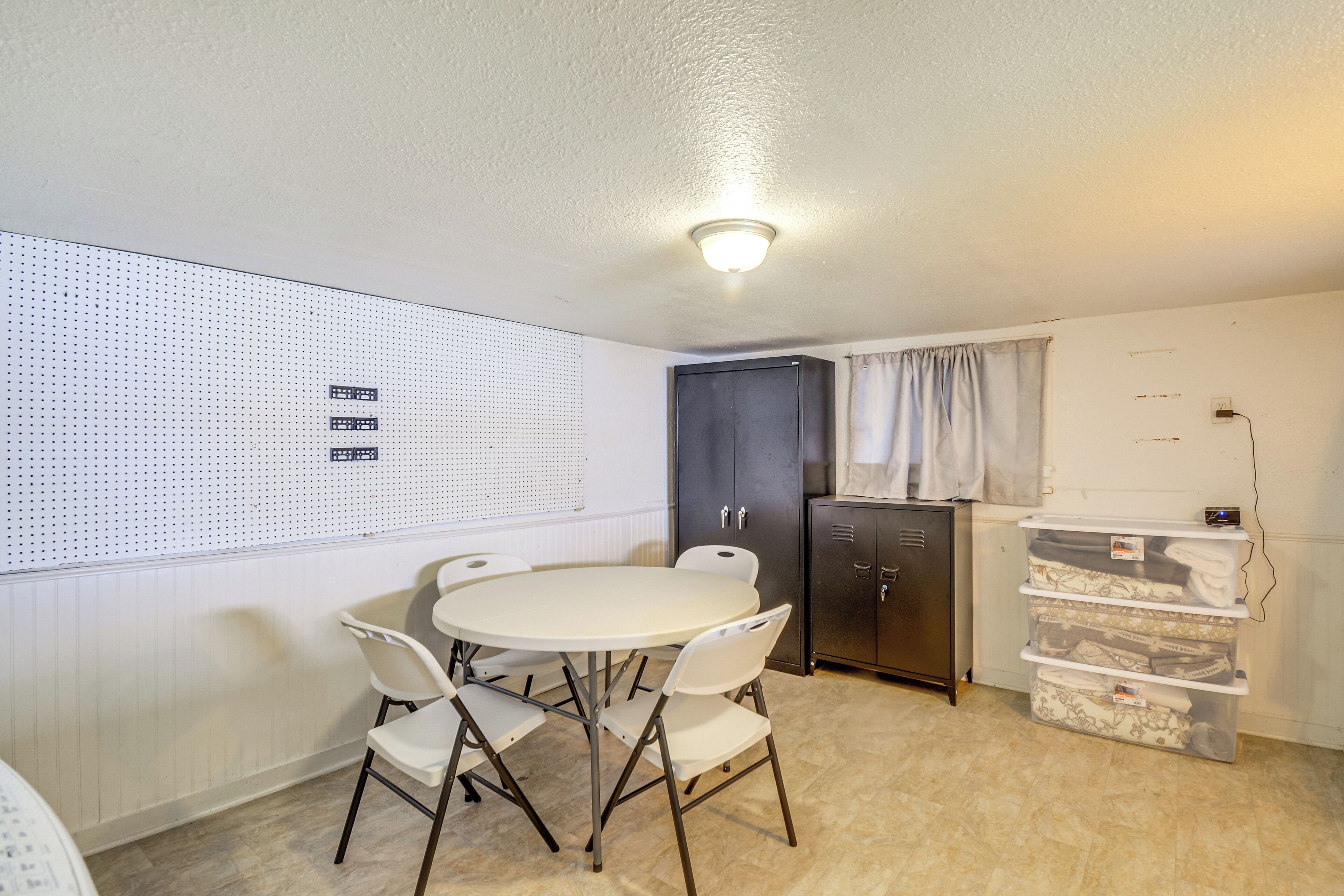 Une pièce de taille modeste, avec une table ronde et quatre chaises, un meuble sombre, un panneau perforé fixé au mur et un meuble de rangement doté de tiroirs.