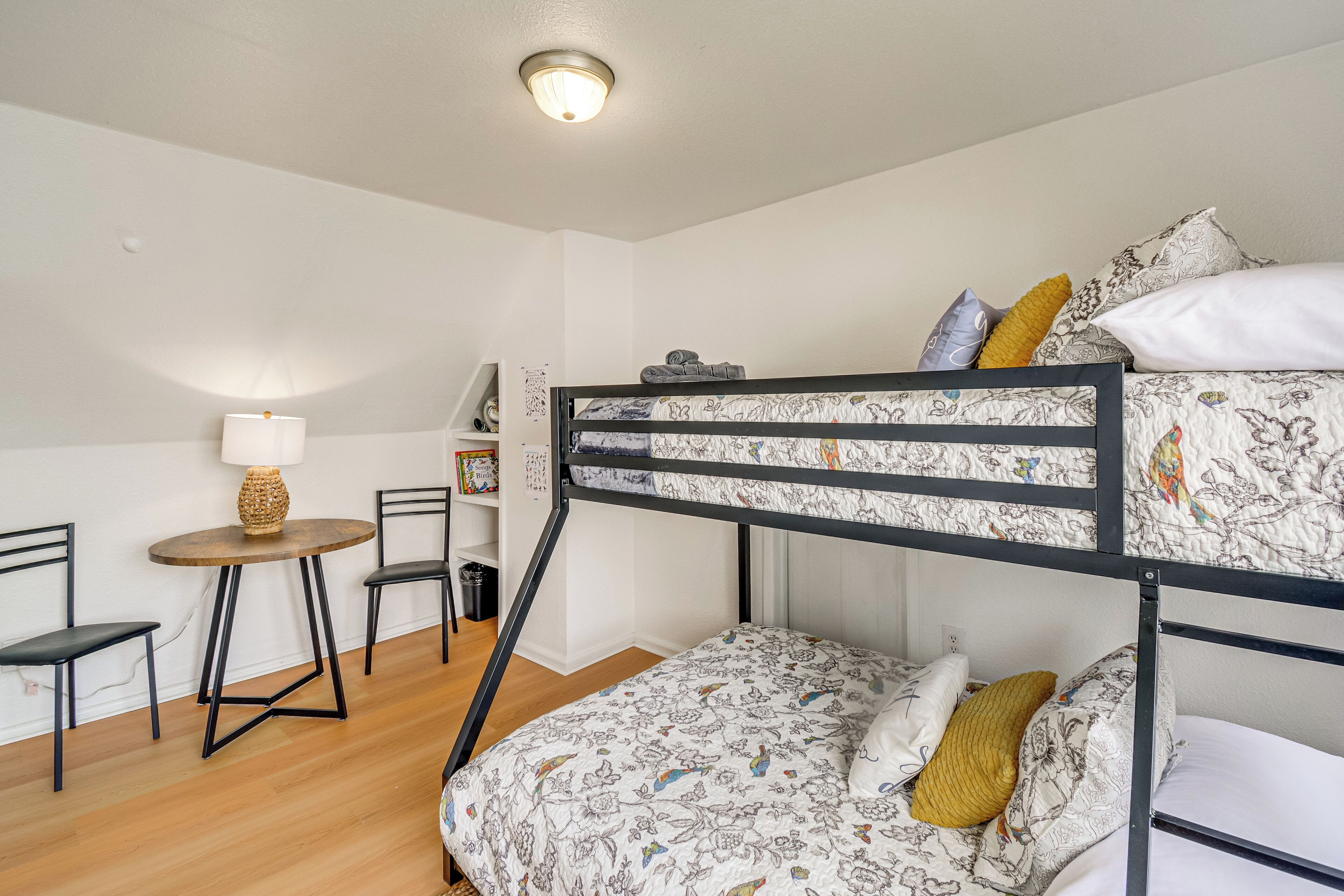 A bunk bed room with a small table and chairs, a lamp, and a shelf with books and decorative items.