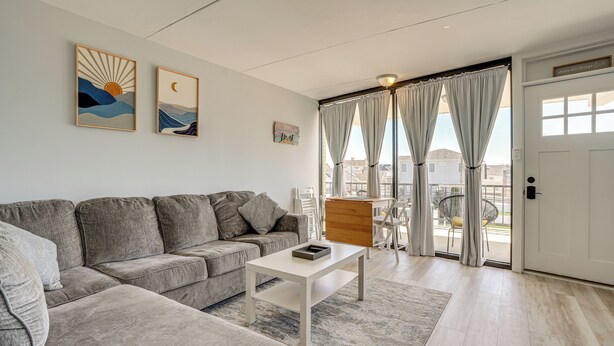 A modern living room with a grey sectional sofa, a white coffee table, and a view of the outdoors through large windows with curtains. - Walk to Beach: North Wildwood Condo w/ Pool Access (North Wildwood)