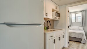 A compact kitchen with white cabinets, a microwave, and a sink. There is a bed with white bedding in the adjacent room. - Walk to Beach: North Wildwood Condo w/ Pool Access (North Wildwood)