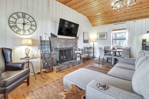A living room with a fireplace, a large wall clock, a flat-screen TV, and a dining area with chairs and a table. - Cozy Cabin + Casita Pinetop Forest Retreat (Pinetop)