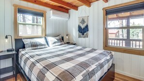 Quarto com colcha xadrez, vigas de madeira no teto, janela com vista e ar-condicionado fixado na parede.