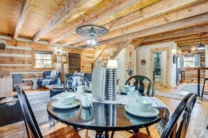 Un interior rústico de madera con zona de comedor que incluye una mesa redonda preparada para cuatro personas, una escalera y una sala de estar con sofá y sillones.