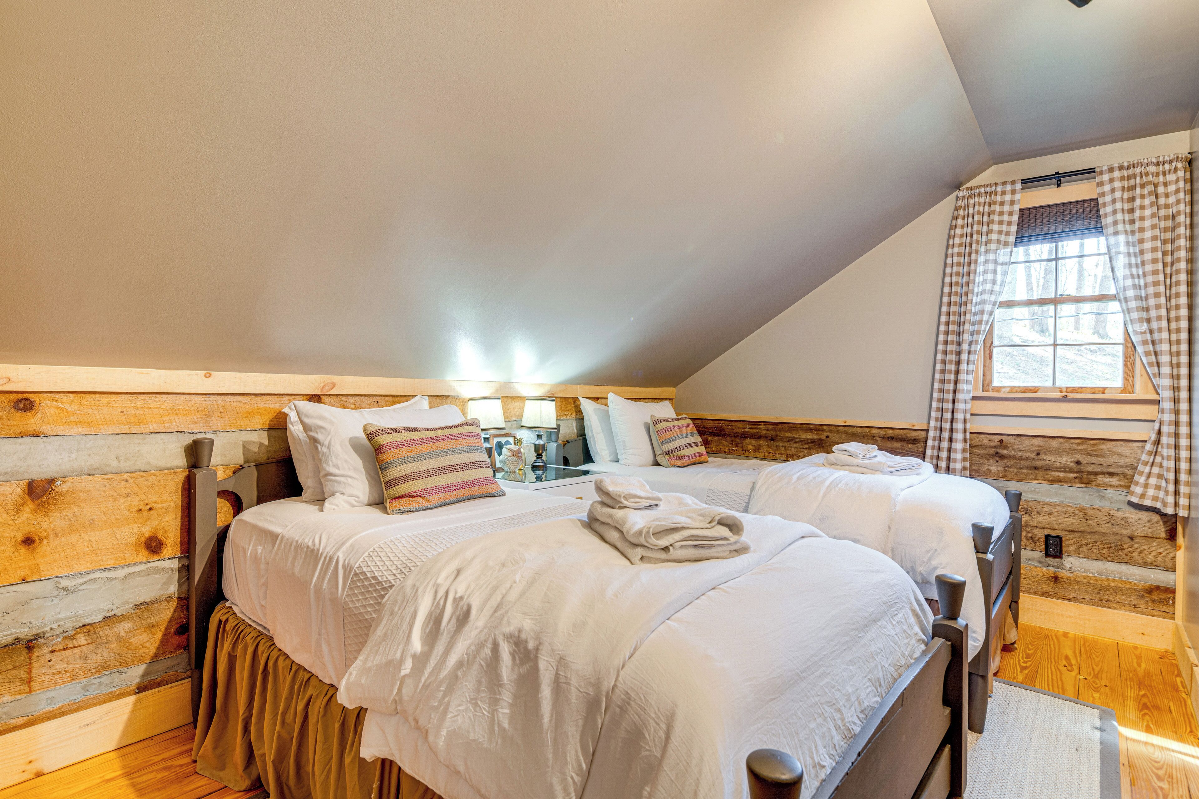 A cozy bedroom with two beds, wooden walls, and a window with checkered curtains.