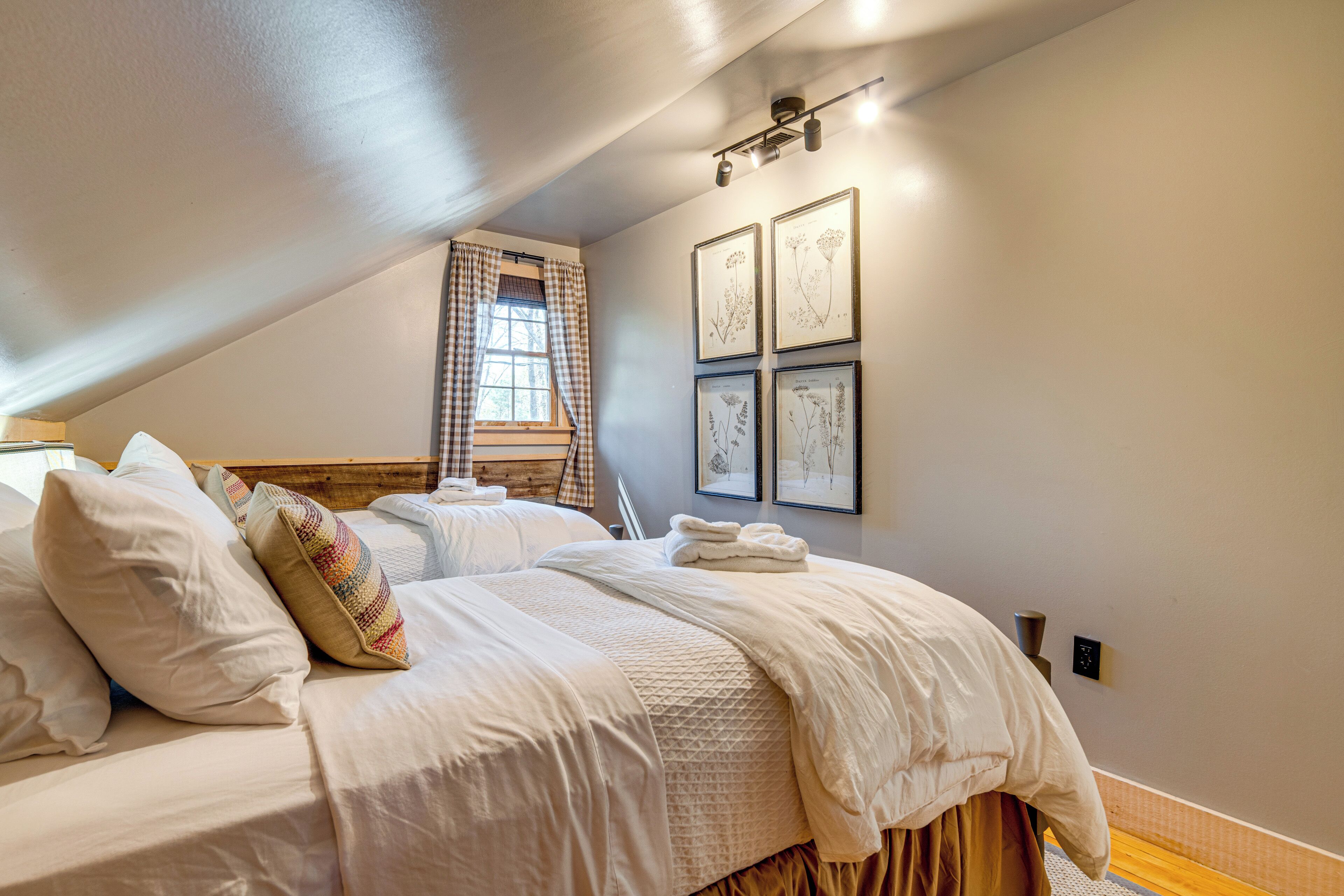 A bedroom with two beds, a window with curtains, and framed botanical prints on the wall.
