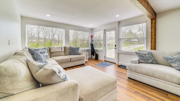 A spacious living room with two beige sofas, a coffee table, and a small side table. Large windows with blinds allow natural light and views of the outdoors. - Peaceful Home w/ Patio on 30 Acres in Coxs Creek! (Coxs Creek)