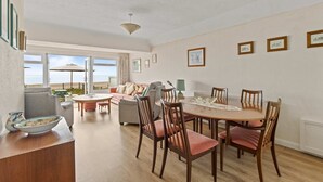 A spacious living area with a dining table, chairs, and a sofa. There are framed pictures on the walls and a view of the beach through large windows. - The Coastal Crab - Family-friendly Three-bed Beach House in Pevensey (Pevensey)