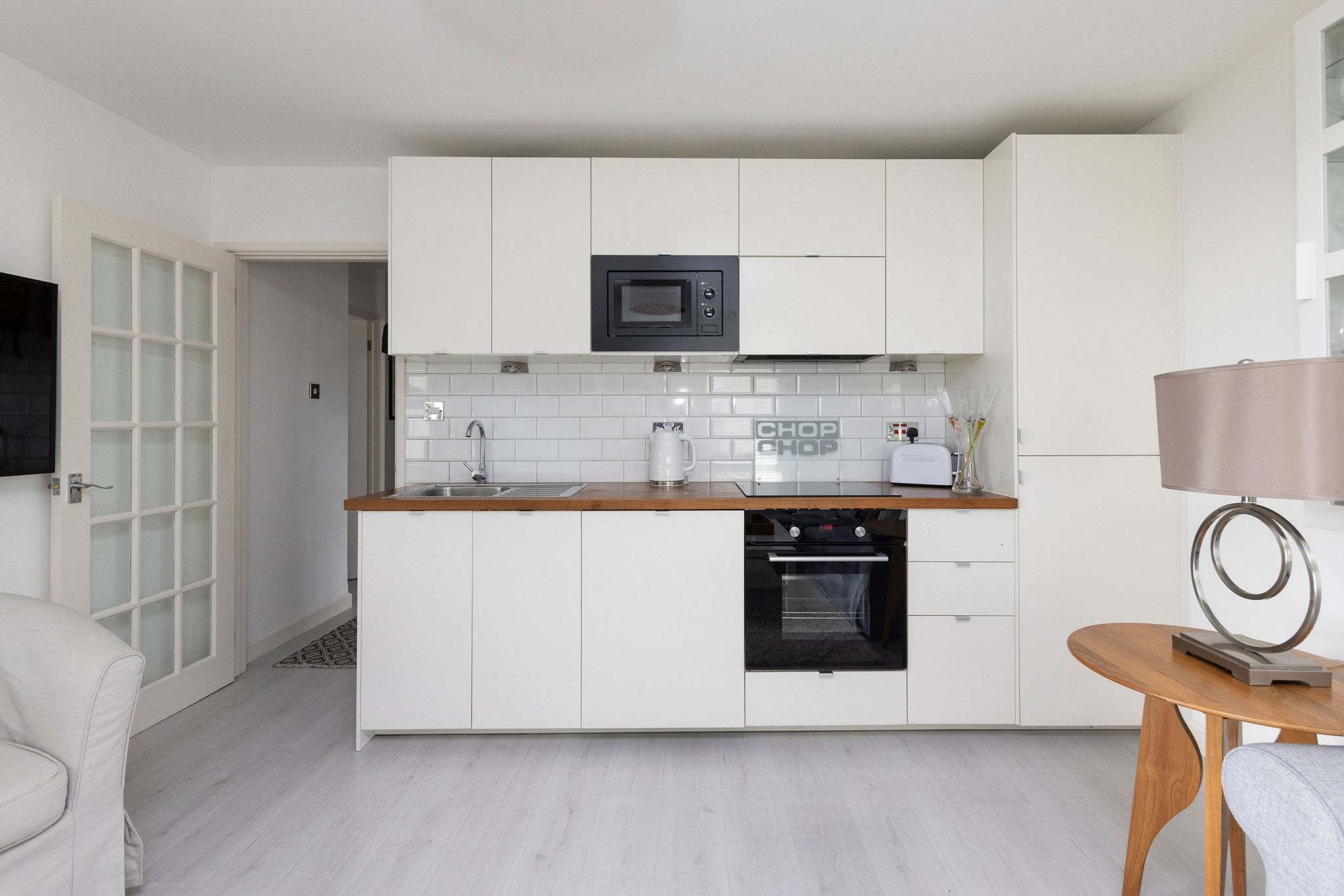 A modern kitchen with white cabinets, a microwave, a coffee machine, and a small oven. There is a wooden table with a lamp and a chair.
