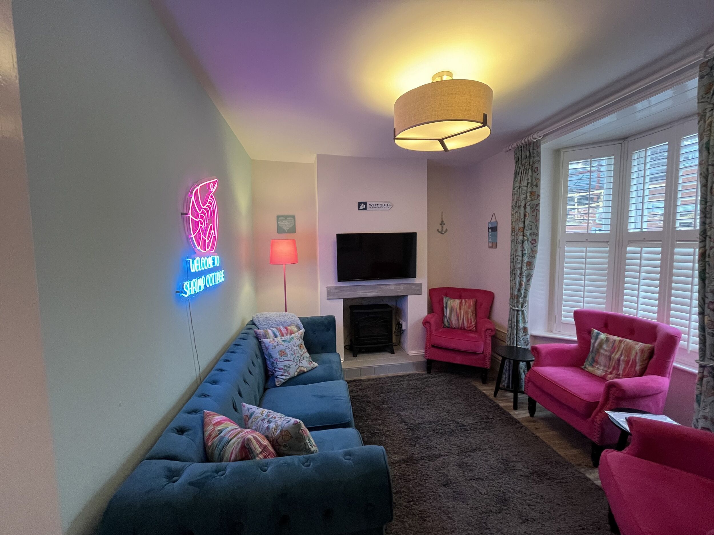 Un salon avec un canapé bleu, un fauteuil rose, un fauteuil rouge, un téléviseur à écran plat, une cheminée et un panneau lumineux au néon.