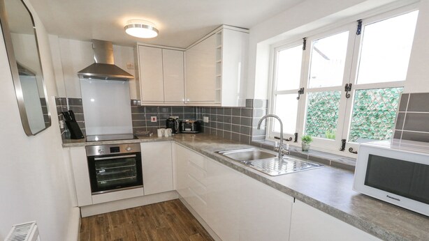 A modern kitchen with white cabinets, a built-in oven, a stainless steel sink, and a window with a view of greenery. - Shrimp Cottage (Weymouth)