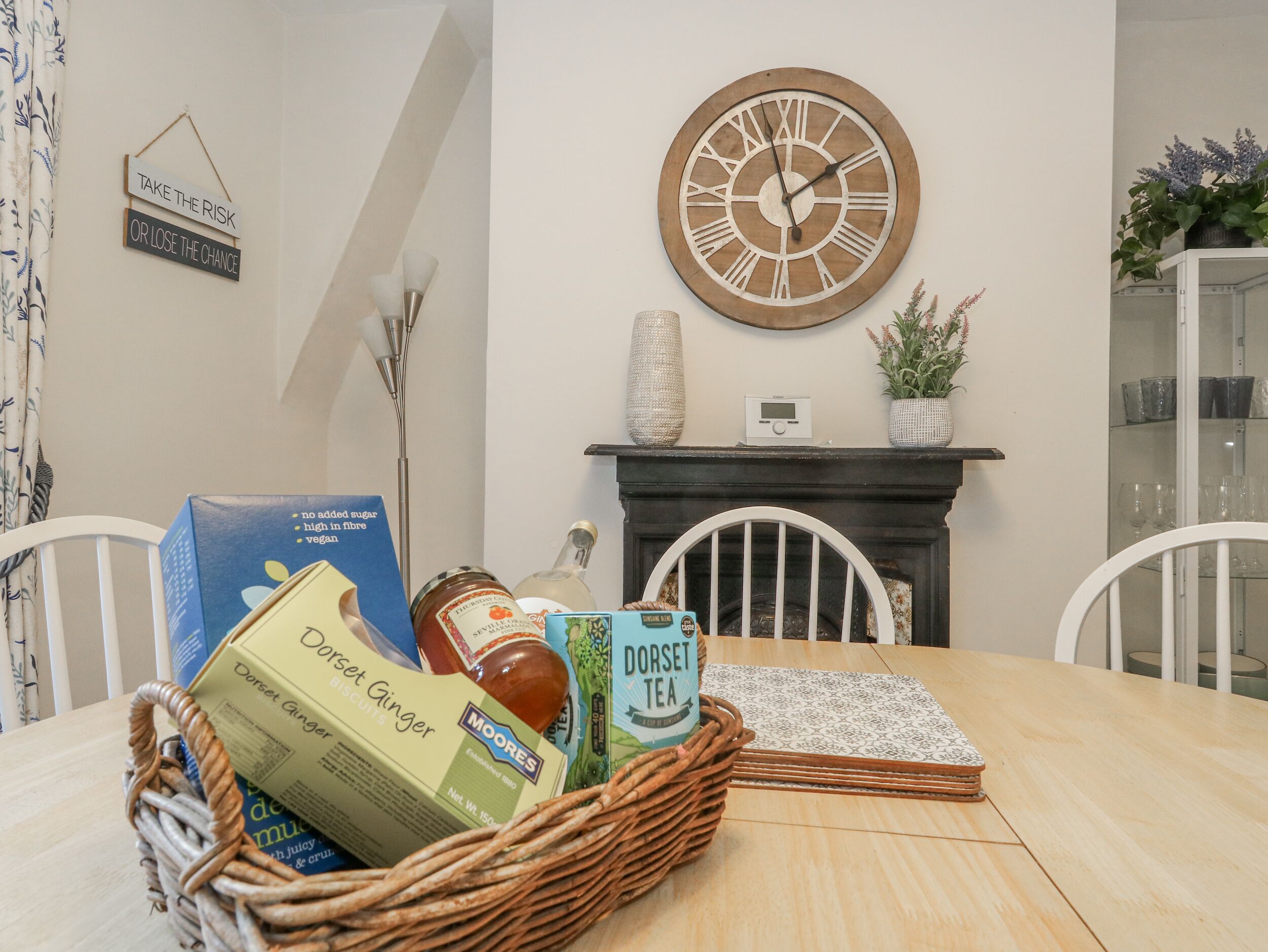 A dining table with a wicker basket containing biscuits, tea, and a jar. A clock and a sign are on the wall.