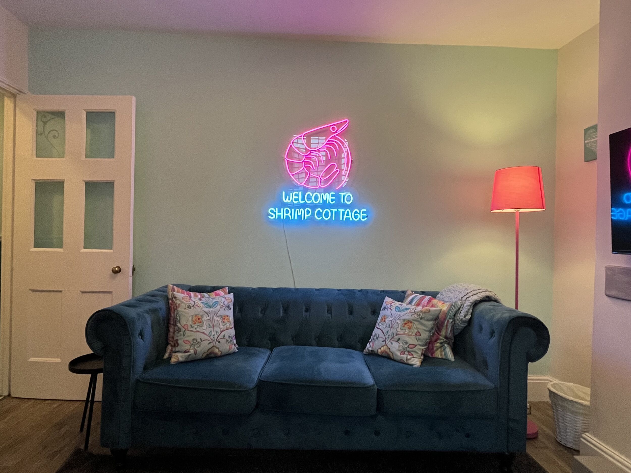 A room with a blue sofa, a neon sign reading "Welcome to Shrimp Cottage," and a red floor lamp.