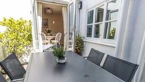 Un área de comedor al aire libre con una mesa y sillas, una planta en maceta y vistas al interior de la casa.