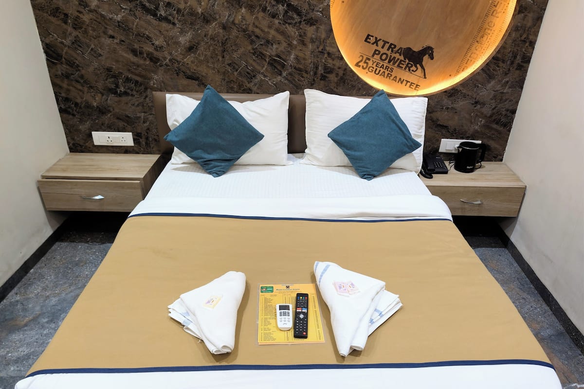 A hotel room with a bed, two pillows, a bedside table, a nightstand, a remote control, and a towel.