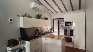 Et lille køkken med hvide skabe, en sort komfur og en mikroovn. Der er en lille bordplade med en elkedel og en plante.