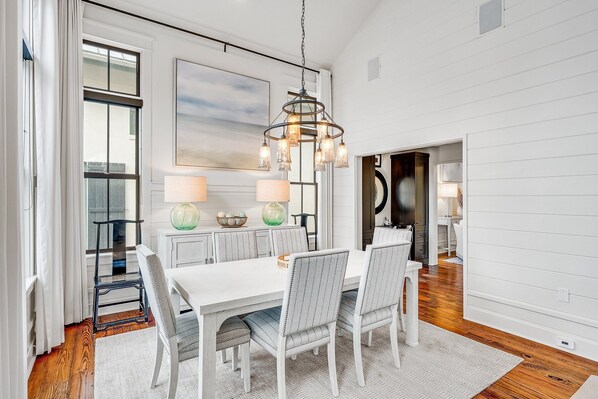 A dining area with a white table and chairs, a chandelier, and a view of the outside. - TreeTop Cottage- Private Pool, Bikes, Steps to the Beach and Town Center. (Rosemary Beach)