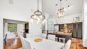A spacious dining area with a large white table, white upholstered chairs, and a wooden floor.
