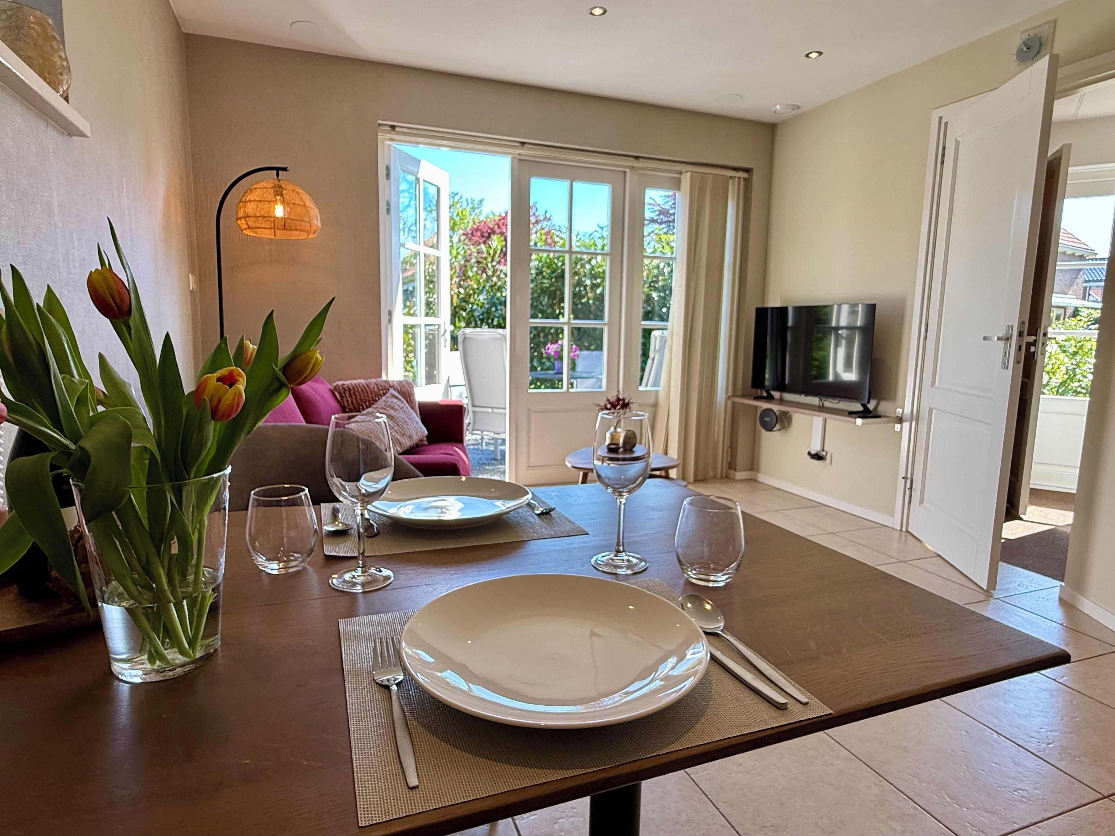A dining area with a table set for two, a vase of tulips, and a television in the background.