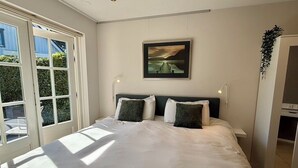 Un dormitorio con una cama grande, dos mesitas de noche, una ventana con persianas, un cuadro enmarcado en la pared y una puerta a la derecha.
