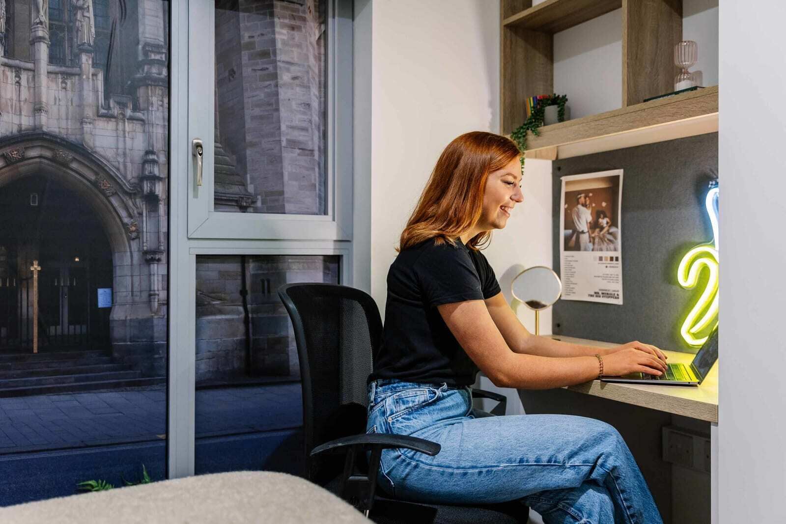 A person working at a desk with a laptop, a neon sign, and a window with a view of a building.