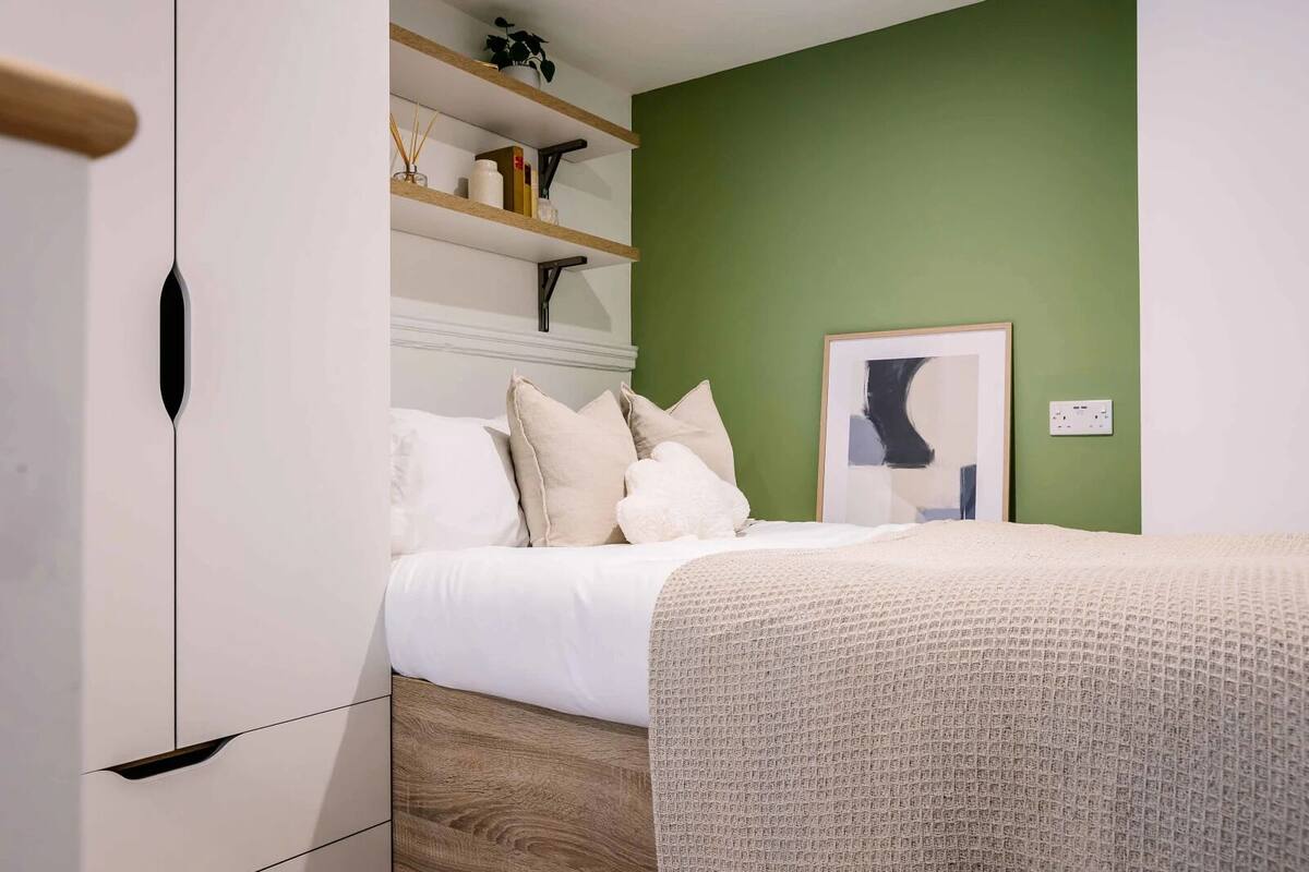 Une chambre à coucher avec un lit, des murs blancs, un mur d’accent vert, un plancher en bois et une étagère avec des livres et une plante.