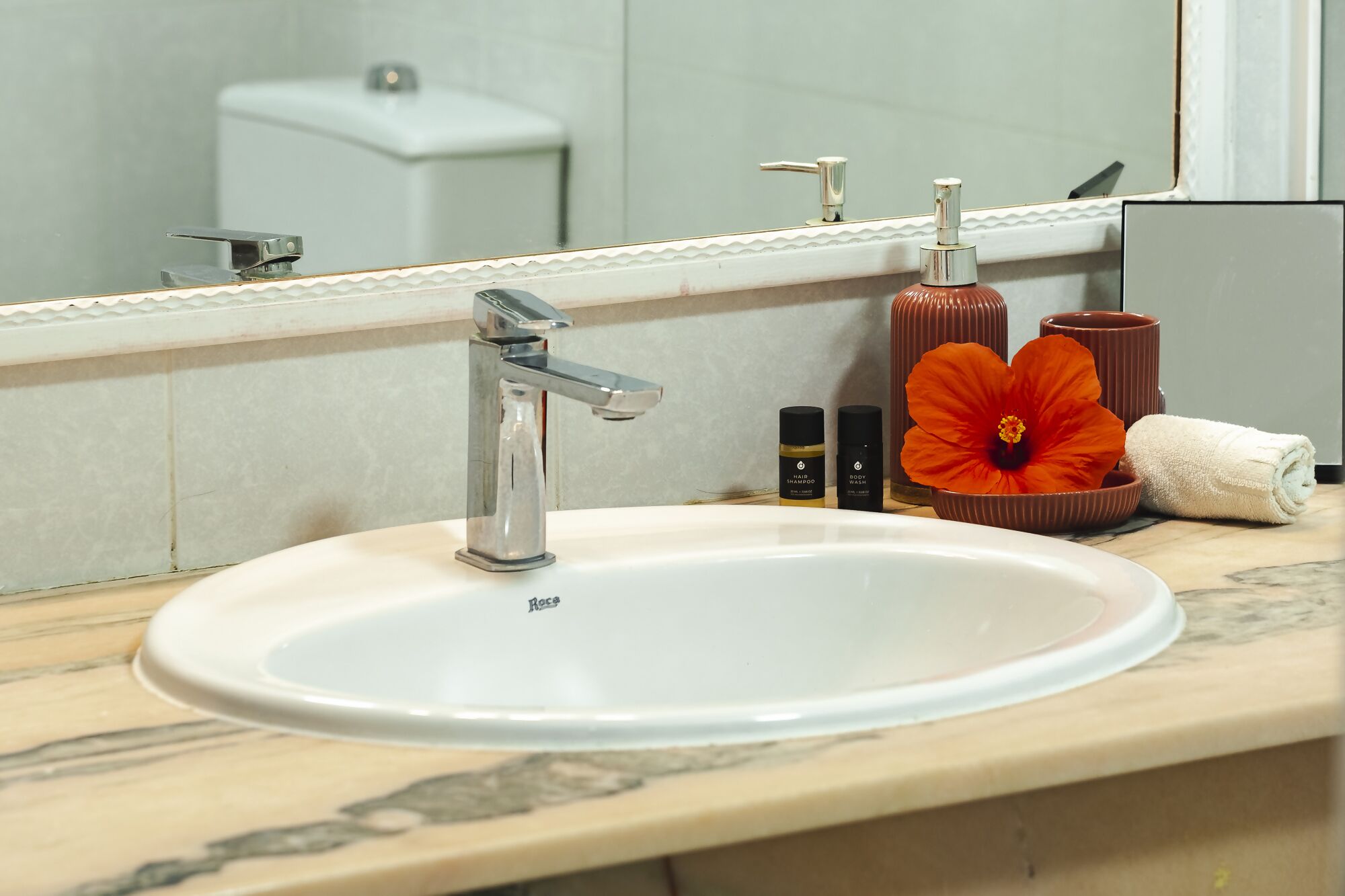 Un bagno con un lavandino bianco, un rubinetto cromato e uno specchio che riflette un water e un asciugamano.