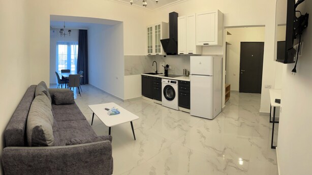Un espacio habitable compacto con un sofá gris, una pequeña mesa blanca, una lavadora y una cocina con armarios blancos y una campana extractora negra.