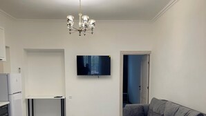 Una sala moderna con un sofá gris, una mesa de centro blanca, un televisor montado en la pared y una zona de cocina con un refrigerador.
