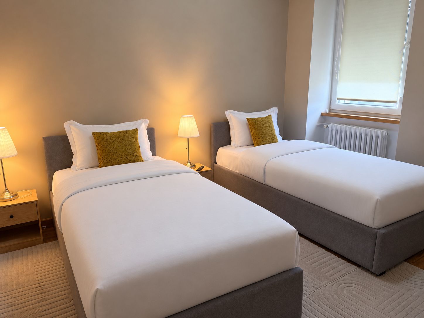 Habitación de hotel con dos camas individuales, mesitas de noche con lámparas, un radiador y una ventana con persianas.