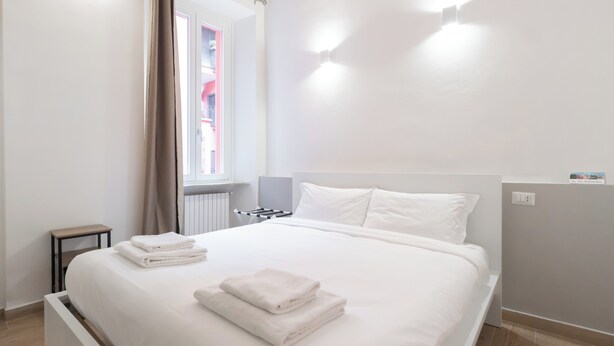A neatly made bed with white linens and towels, a small bedside table, a window with curtains, and a wall-mounted light fixture. - Italianway - Frisi 8 B (Milan)