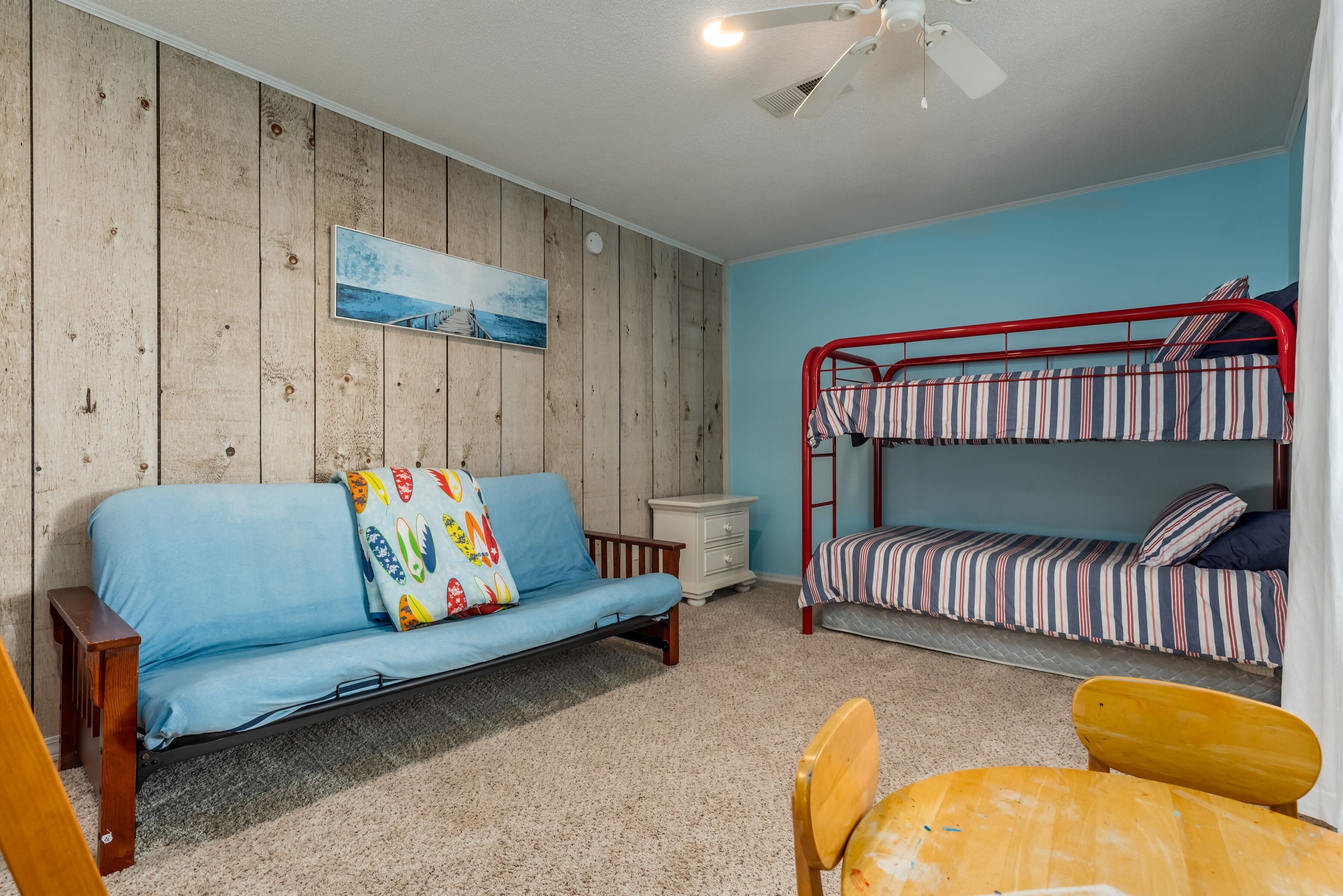 Una habitación con pared de madera, un sofá azul, una cama litera, una mesita y un ventilador de techo.