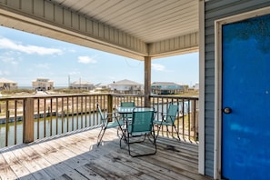 A wooden deck with a covered area, a blue door, and a view of houses across a body of water. - Anchor Eways - 2235 Mallard St 3 Bedroom Home (Dauphin Island)