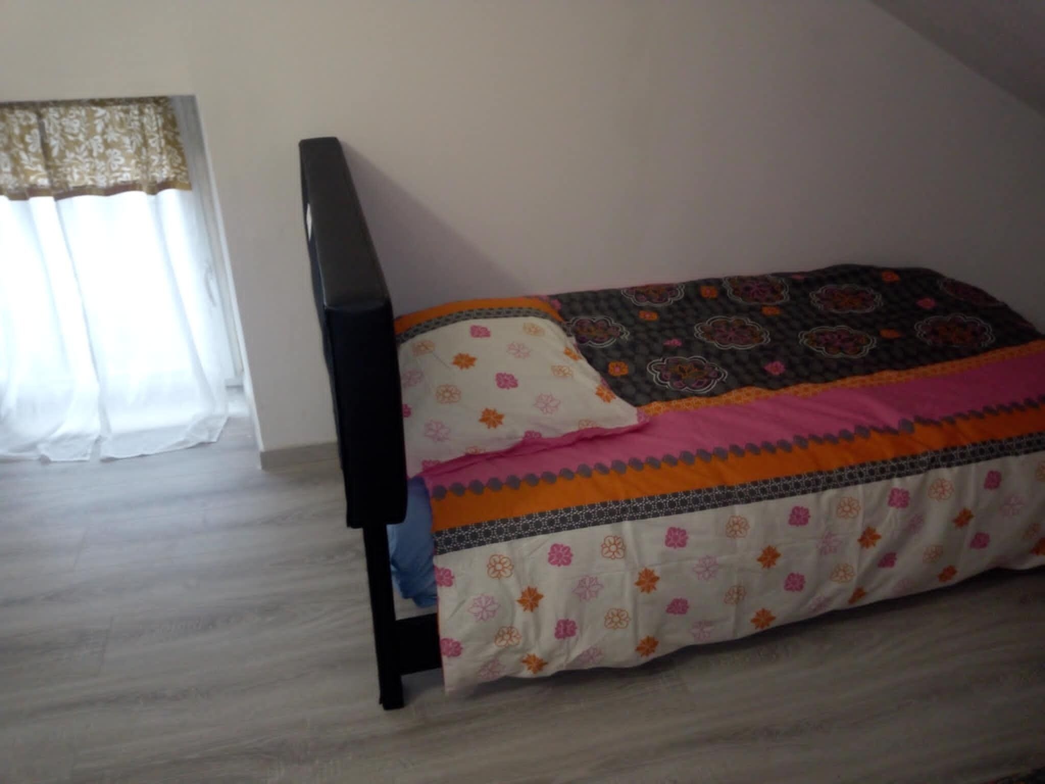 A bedroom with a bed, a window with sheer curtains, and a wooden floor.