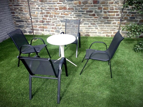 Un patio con una mesa redonda y cuatro sillas, rodeado de césped artificial, frente a un muro de piedra.