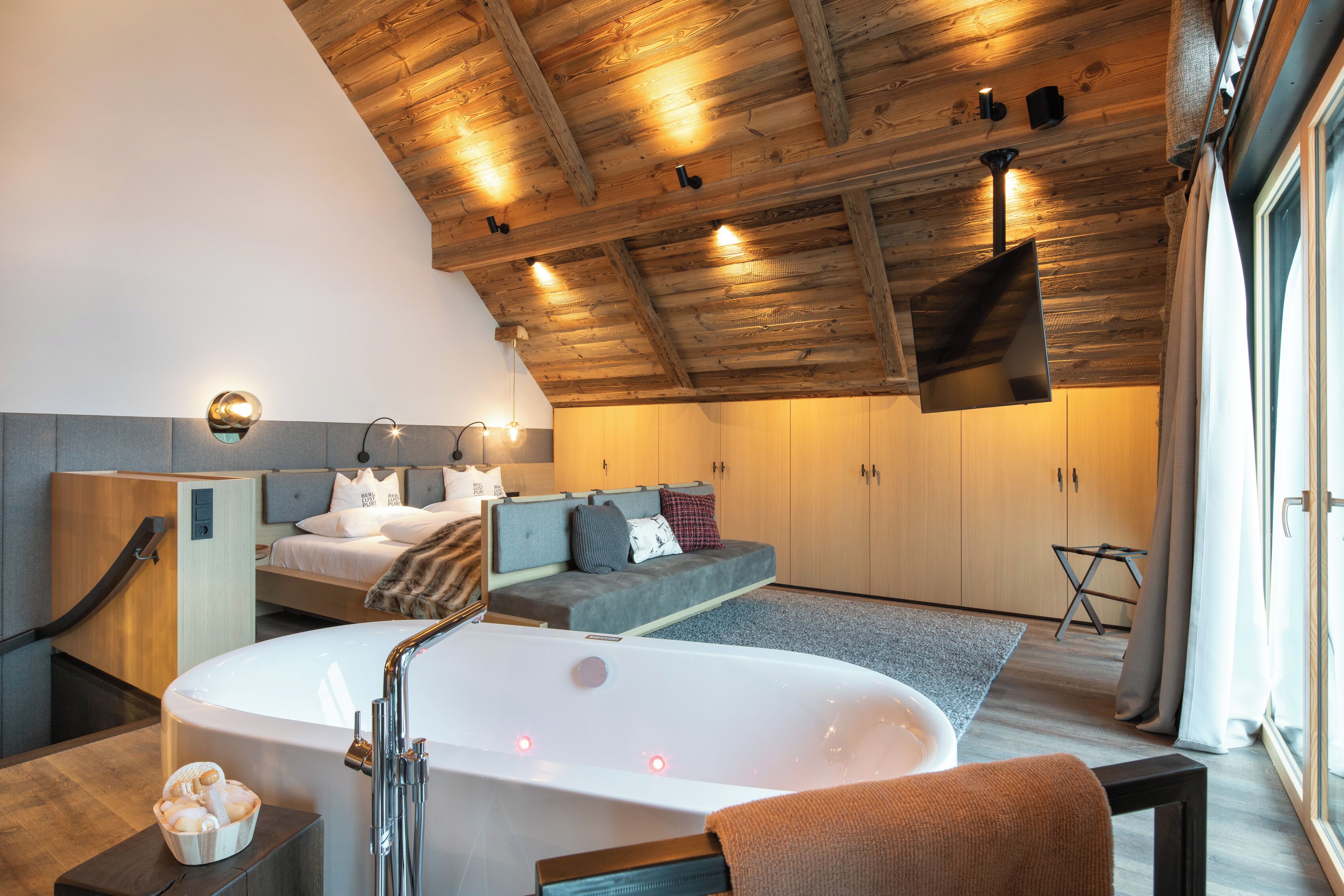 A modern bedroom with a large bathtub, wooden ceiling beams, a sofa, and a bed with pillows.