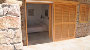 Een stenen muur met een houten deur, leidend naar een slaapkamer met een bed en nachtkastje.