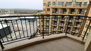 Un balcón con vista a un edificio de varios pisos, una zona pavimentada con coches aparcados y un paisaje distante.