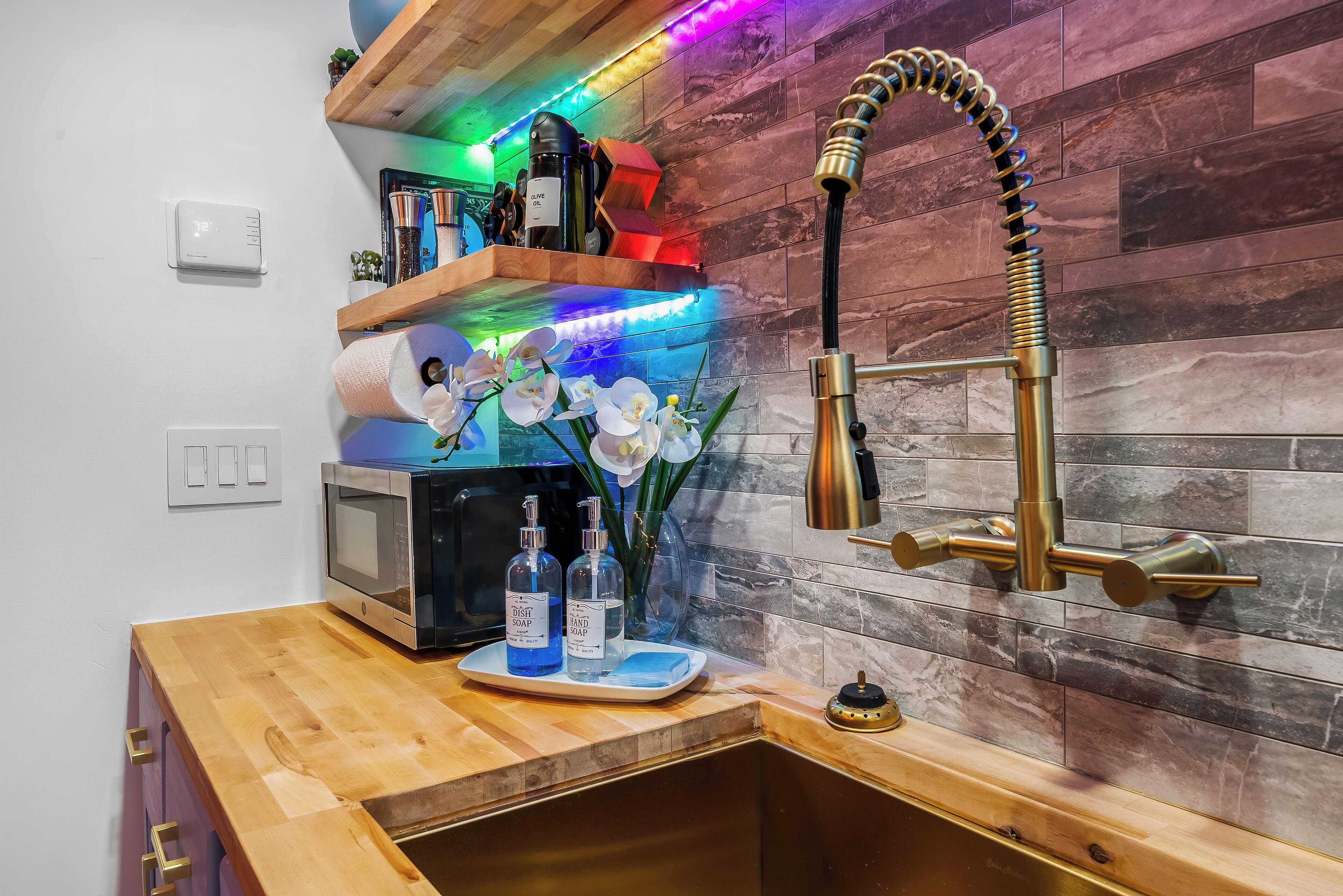 A modern kitchen with a wooden countertop, a built-in microwave, and a stainless steel sink. There are two bottles of soap on the counter and a vase with white flowers.