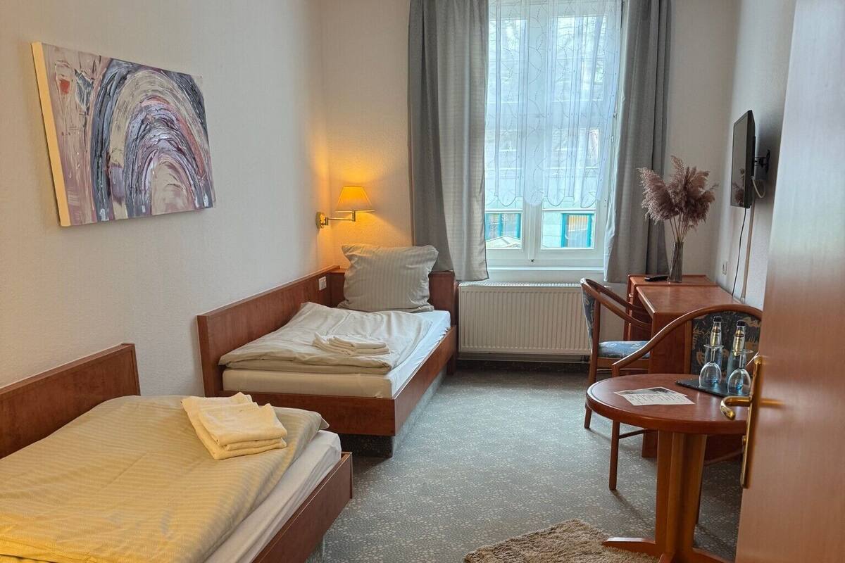 Ein Hotelzimmer mit zwei Einzelbetten, einem Holztisch mit Stuhl, einem Heizkörper und einem Fenster mit Vorhängen.