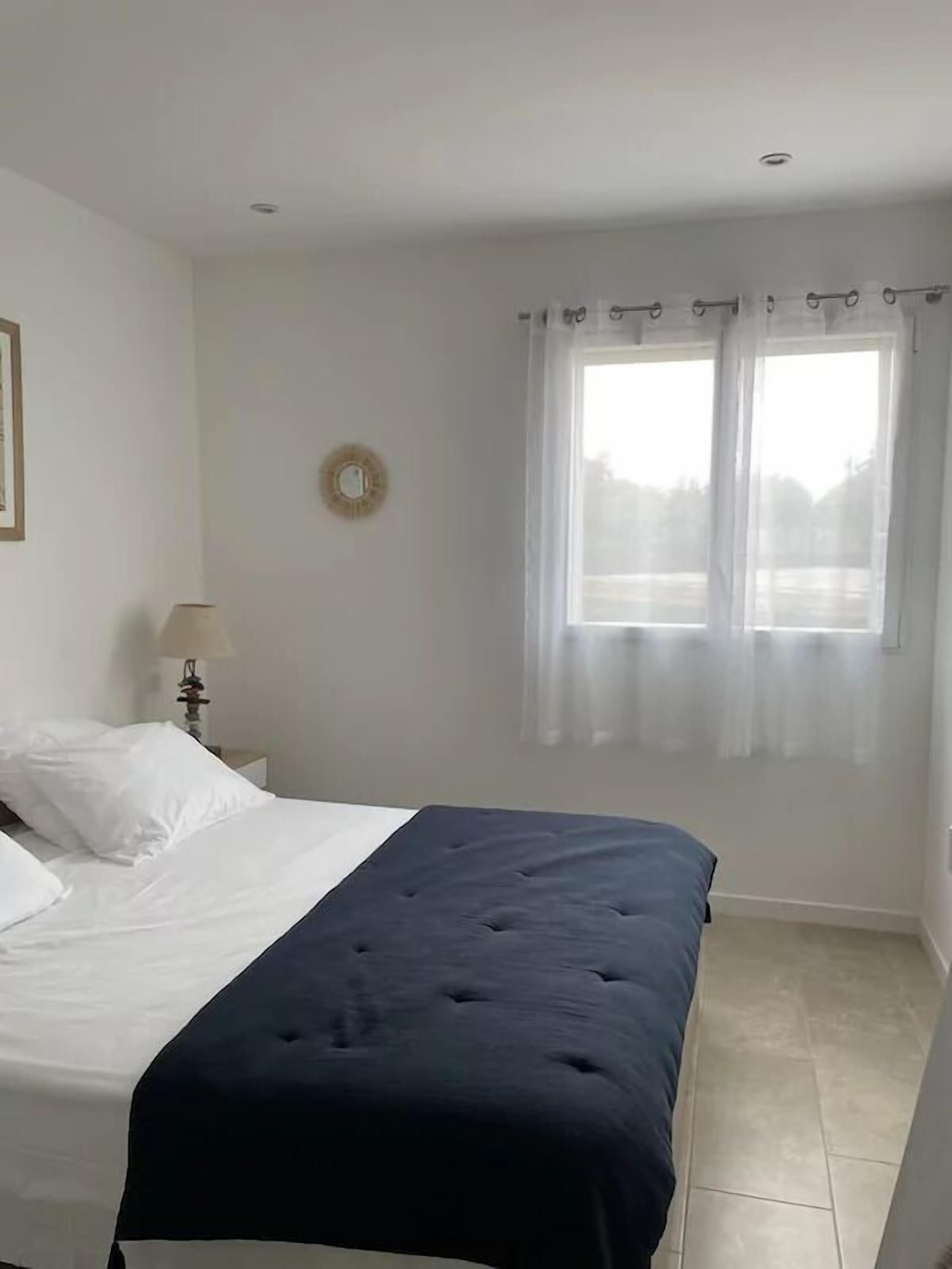 A bedroom with a bed, a nightstand, a lamp, a window with curtains, and a wall clock.