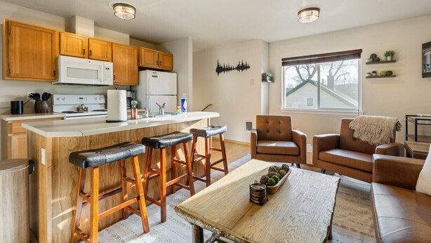 A modern kitchen with wooden cabinets, a white stove, and a bar area with stools. There is a dining table with a vase and a candle. The room has a large window and a brown leather sofa. - Downtown Kalispell Condo Near Glacier & Whitefish (Kalispell)