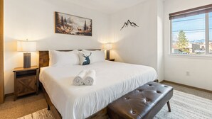 A hotel room with a large bed, two bedside lamps, a ceiling fan, a mountain painting, a mountain silhouette decoration, and a window with a view of a tree and buildings. - Downtown Kalispell Condo Near Glacier & Whitefish (Kalispell)
