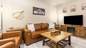 A living room with a brown leather sofa, a wooden coffee table, a flat-screen TV, and wall-mounted artwork. - Downtown Kalispell Condo Near Glacier & Whitefish (Kalispell)
