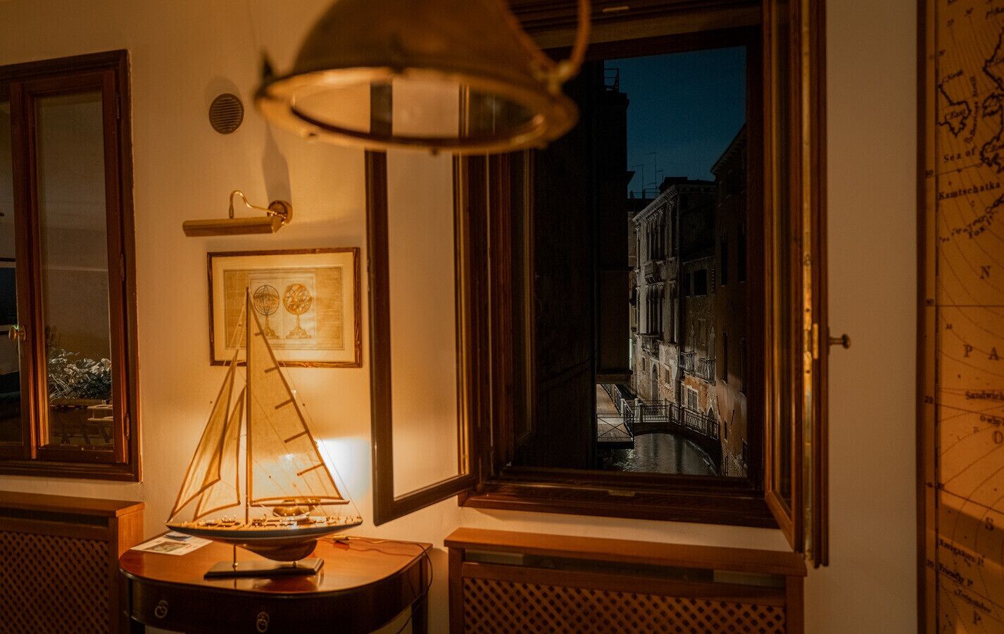 A room with a large window overlooking a canal, a map on the wall, and a table with a lamp and a model ship.