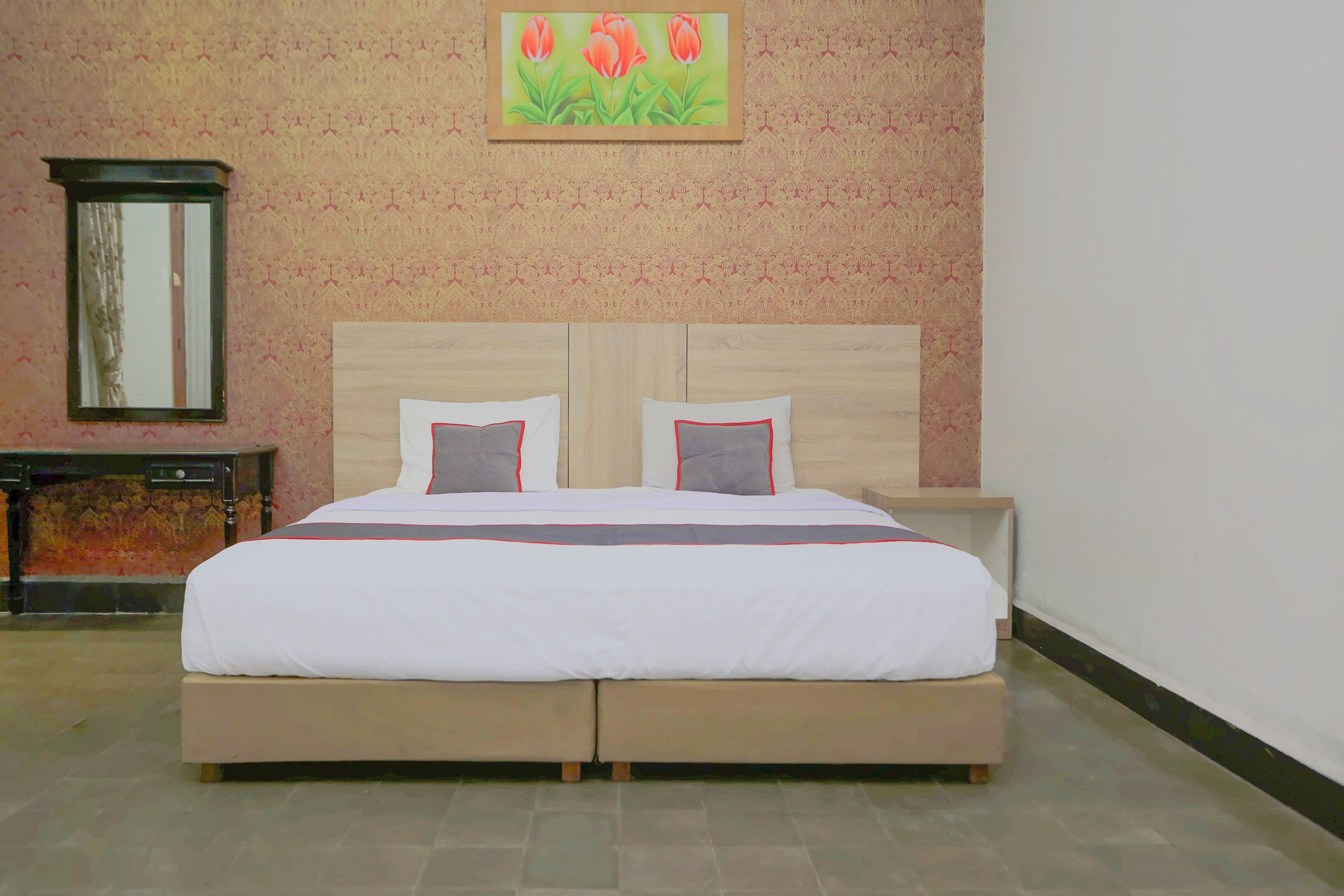 A double bed with a wooden headboard and two pillows, a nightstand, a mirror, and a painting of flowers on the wall.