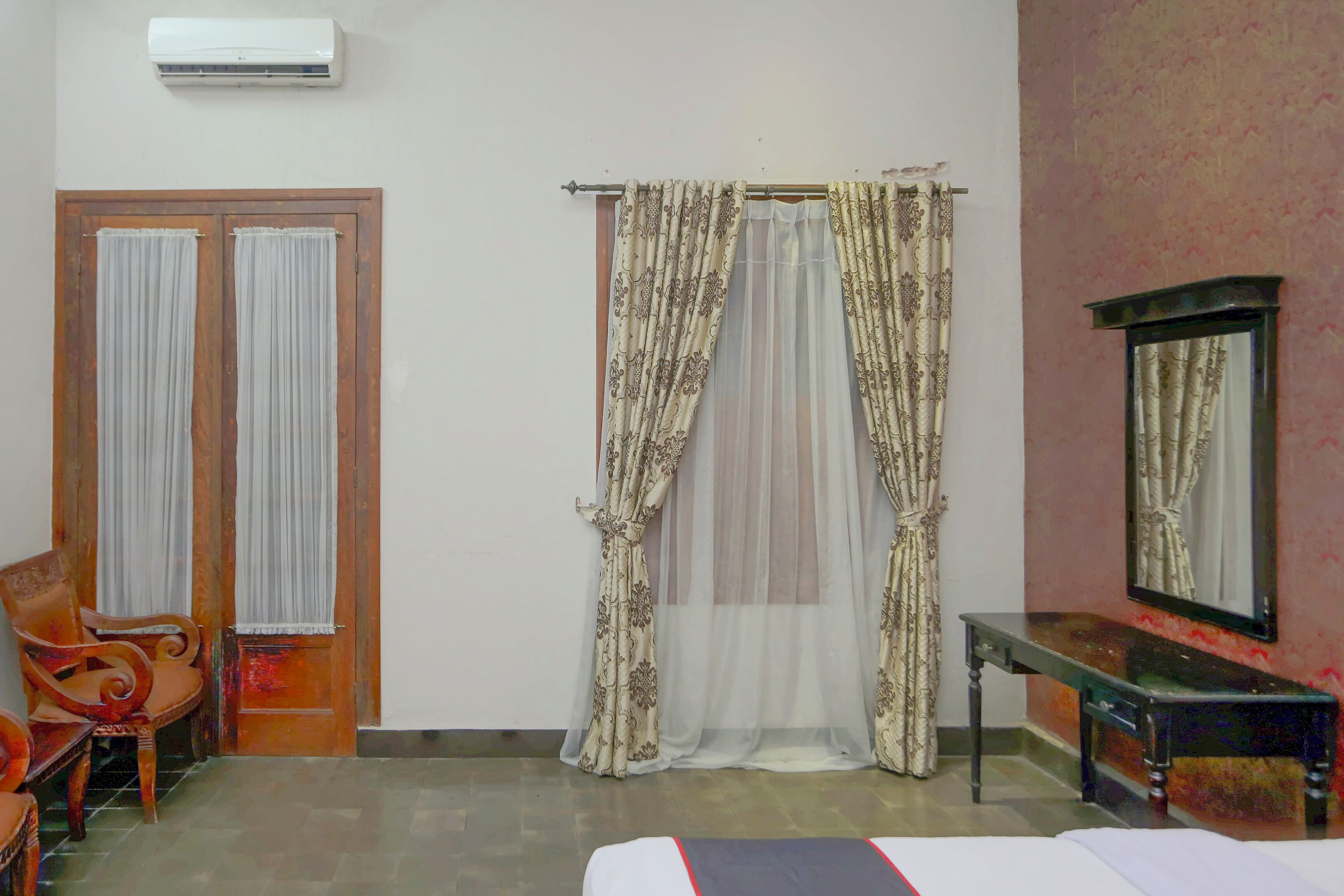 A room with a bed, a wooden chair, a small table, and a mirror. There is an air conditioning unit on the wall and a window with curtains.