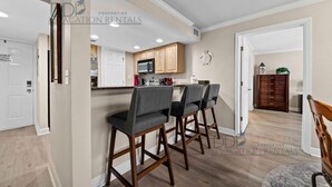 A modern kitchen with wooden cabinets, a microwave, and a built-in oven. There are two bar stools and a dining table in the adjacent area.